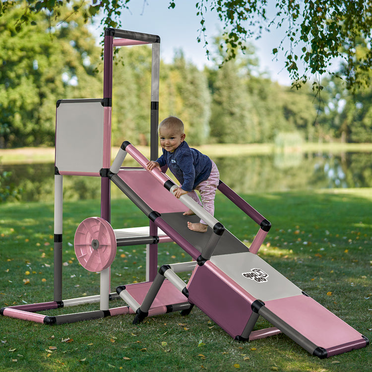 Małe dziecko boso patrzy w aparat z rampy QUADRO Evolution Berry, koło wielofunkcyjne po lewej stronie wieży do zabawy